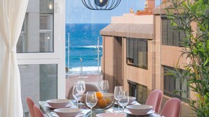 A dining table set with pink plates, glasses, and a fruit bowl, overlooking a beach with buildings in the background. - Deluxe Beach Canteras Flat 3br Wifi Free 1ºA (Palmas De Gran Canaria, Las)