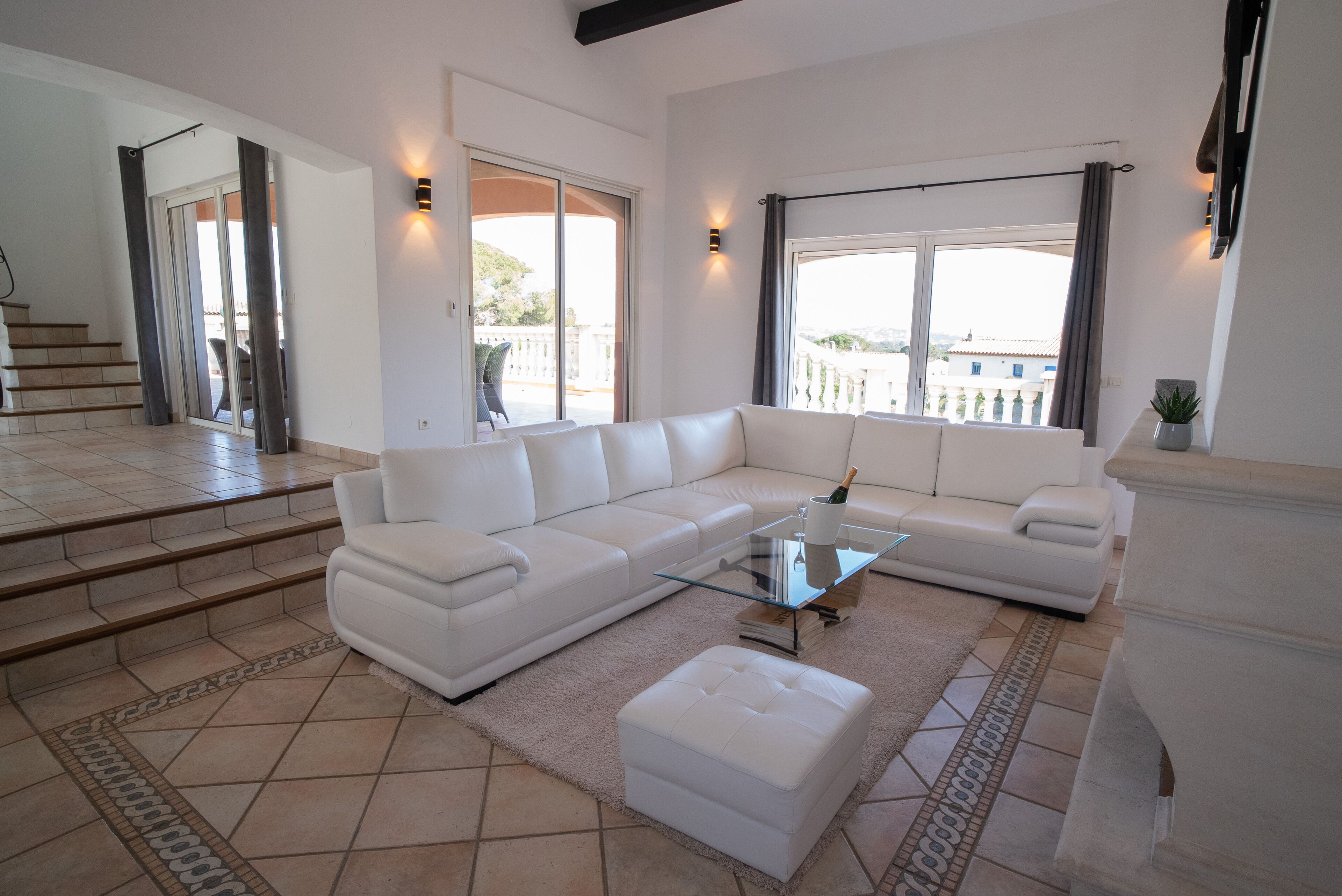 A spacious living room with a white sectional sofa, a glass coffee table, and a small ottoman. There is a staircase leading to an outdoor area with a view of buildings and greenery.
