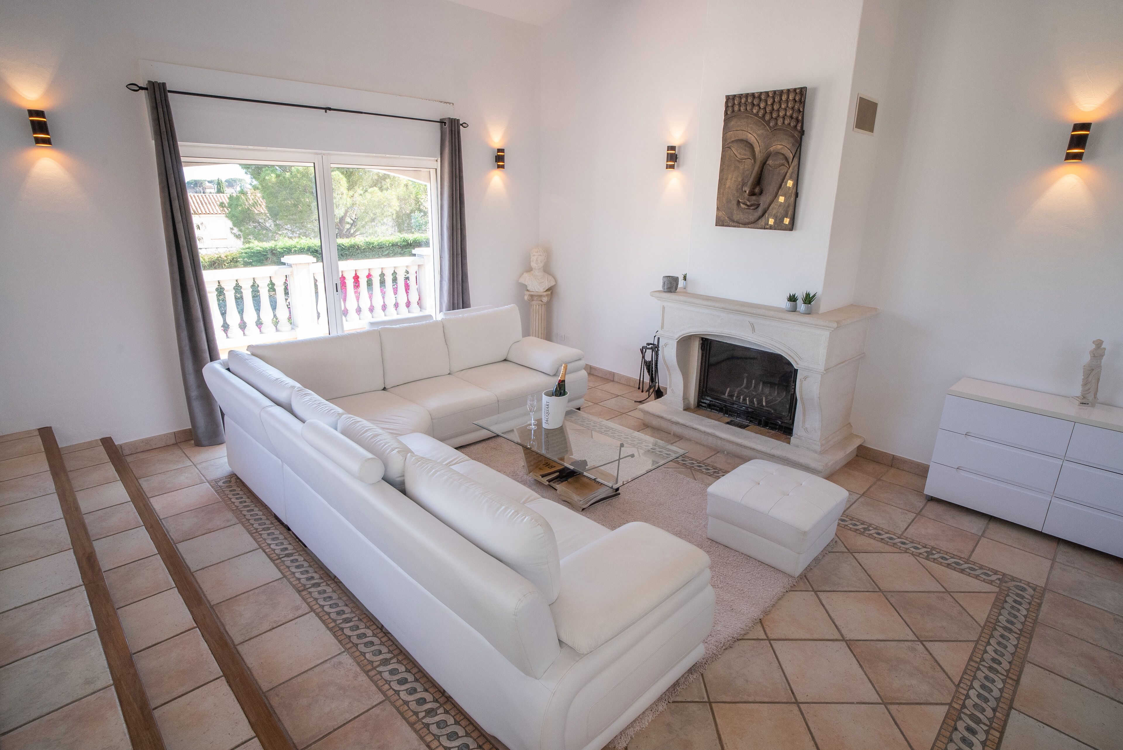 A modern living room with a white sofa, a glass coffee table, a fireplace, and a view of the outside balcony.