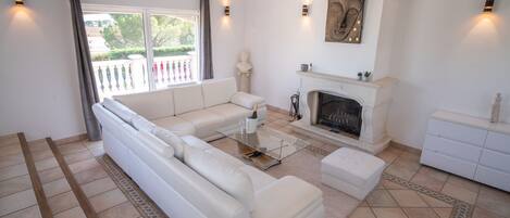 A modern living room with a white sofa, a glass coffee table, a fireplace, and a view of the outside balcony.