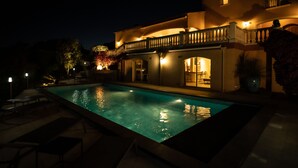 Ein Nachtbild eines zweistöckigen Hauses mit Swimmingpool, Außenbeleuchtung und einem Balkon.