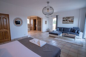 Une chambre moderne avec un lit, un canapé et une table basse. On aperçoit une salle de bain avec une douche et des toilettes à travers une arche.