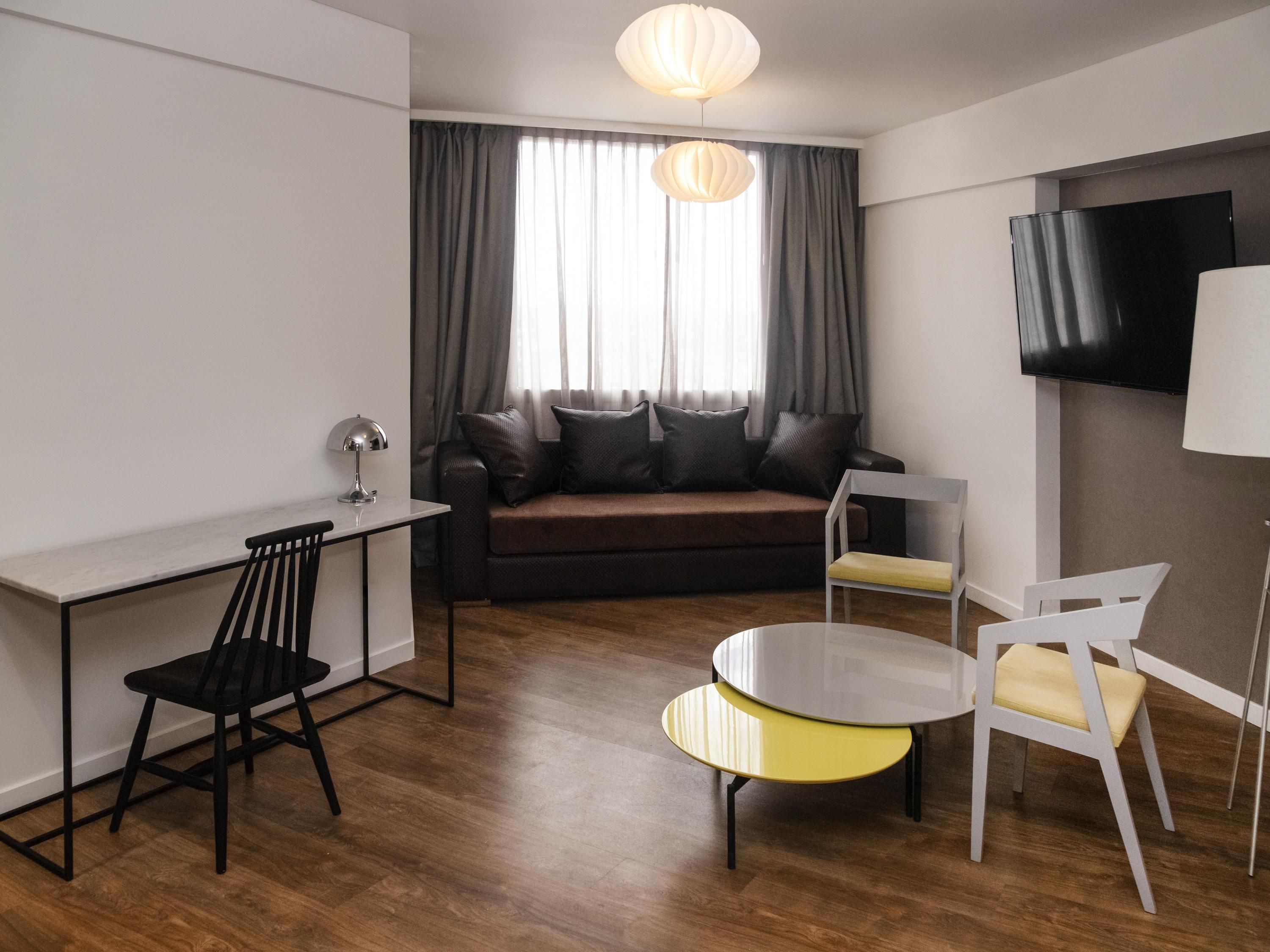 A modern living room with a sofa, a desk, a chair, a small table, and a TV mounted on the wall.