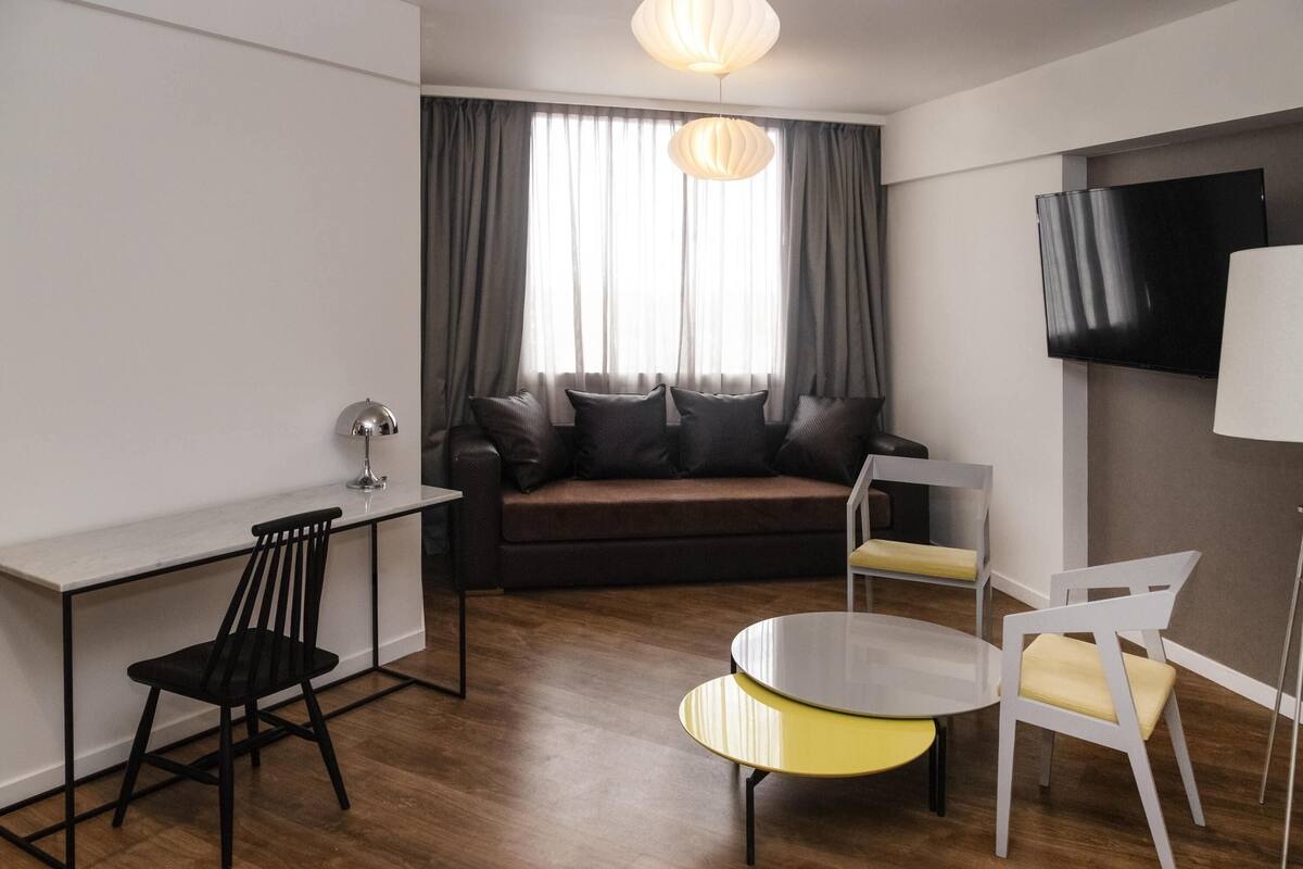 Een moderne woonkamer met een bank, een bureau, een stoel, een bijzettafeltje en een tv aan de muur.