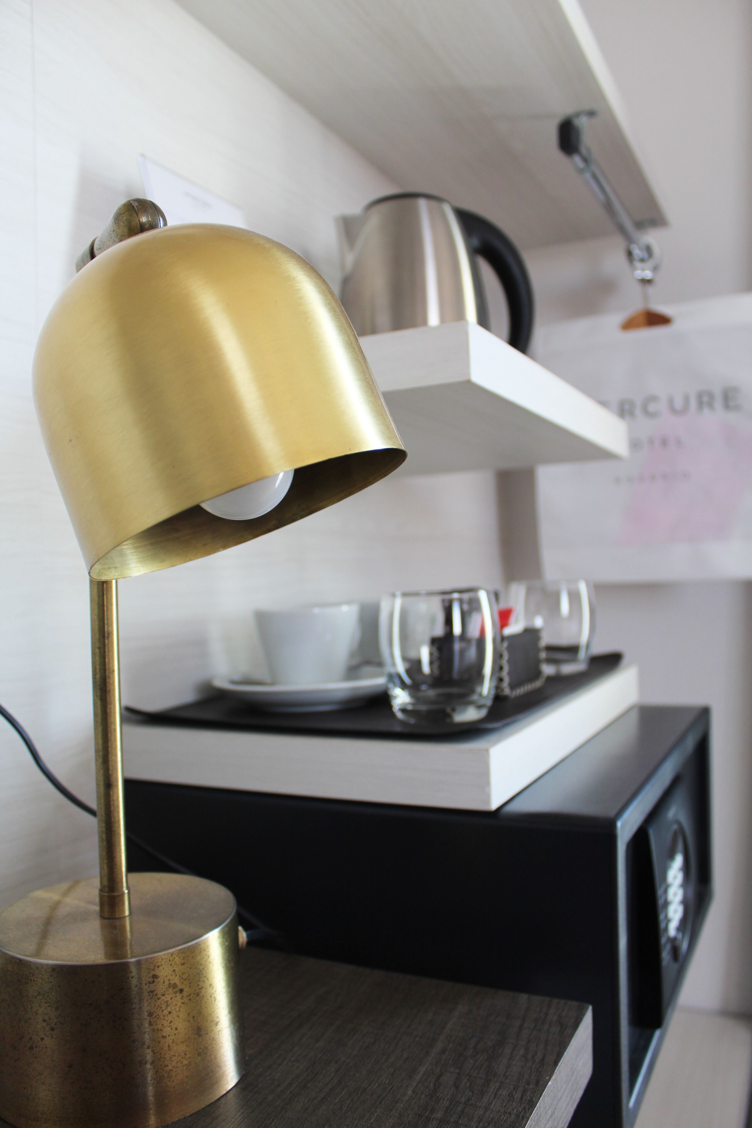 A desk lamp on a desk with a black surface, a white shelf with a white cup and saucer, and a black cabinet with a white label.