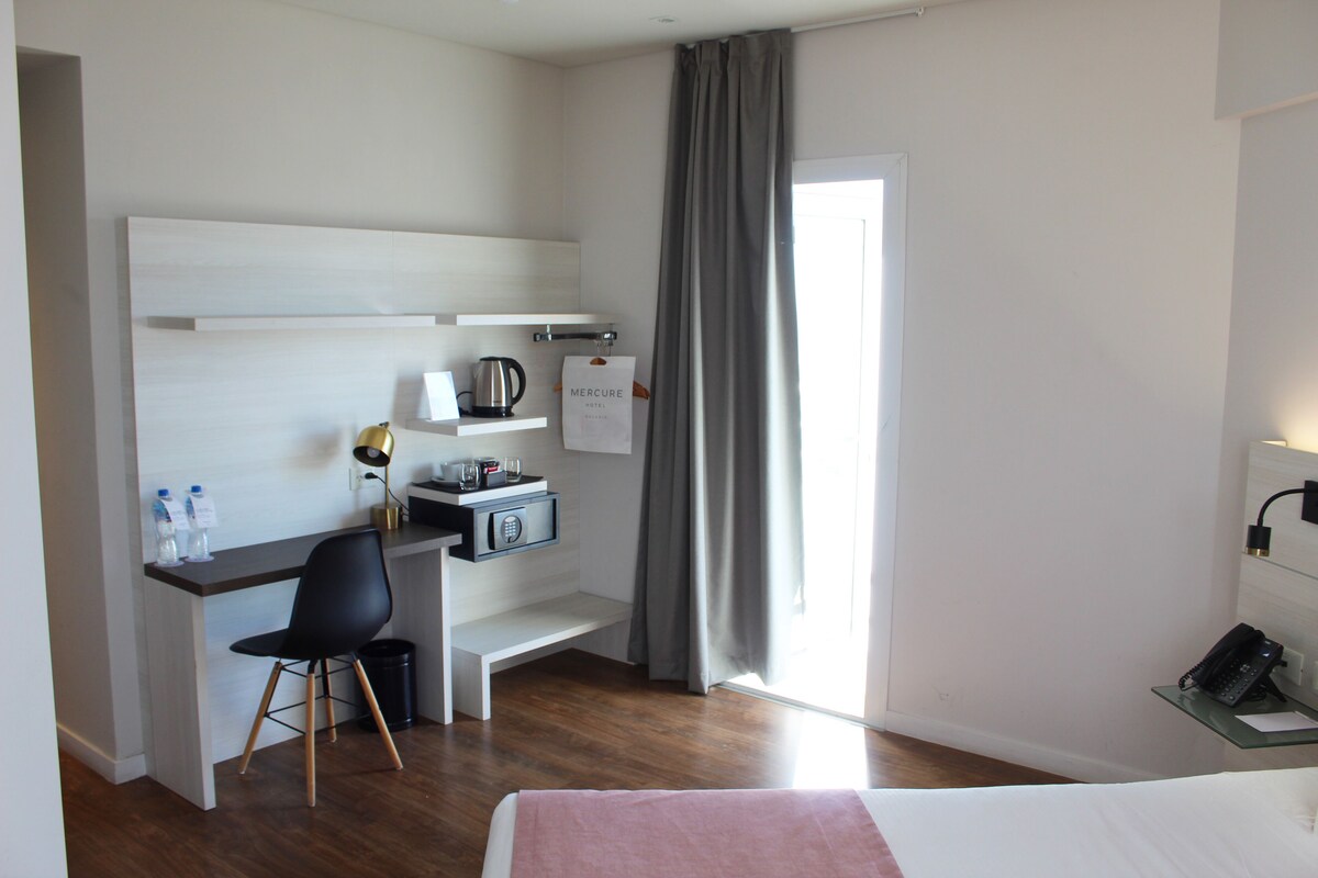Hotelkamer met een bed, bureau met stoel, koffiezetapparaat, kluisje, prullenbak en telefoon.