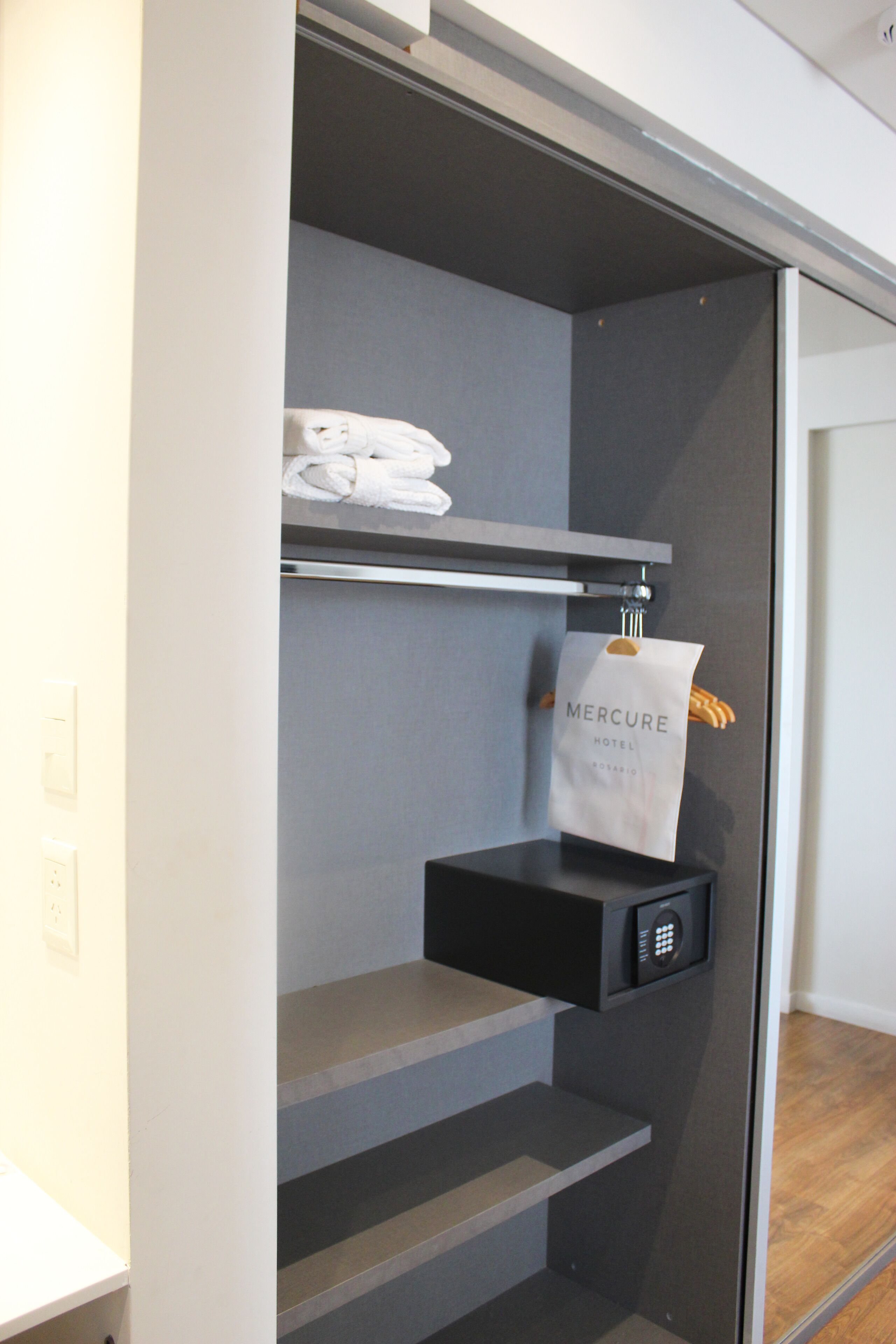 Habitación de hotel con puerta corredera, un perchero con toallas dobladas y un bolso del hotel Mercure colgado en la puerta.