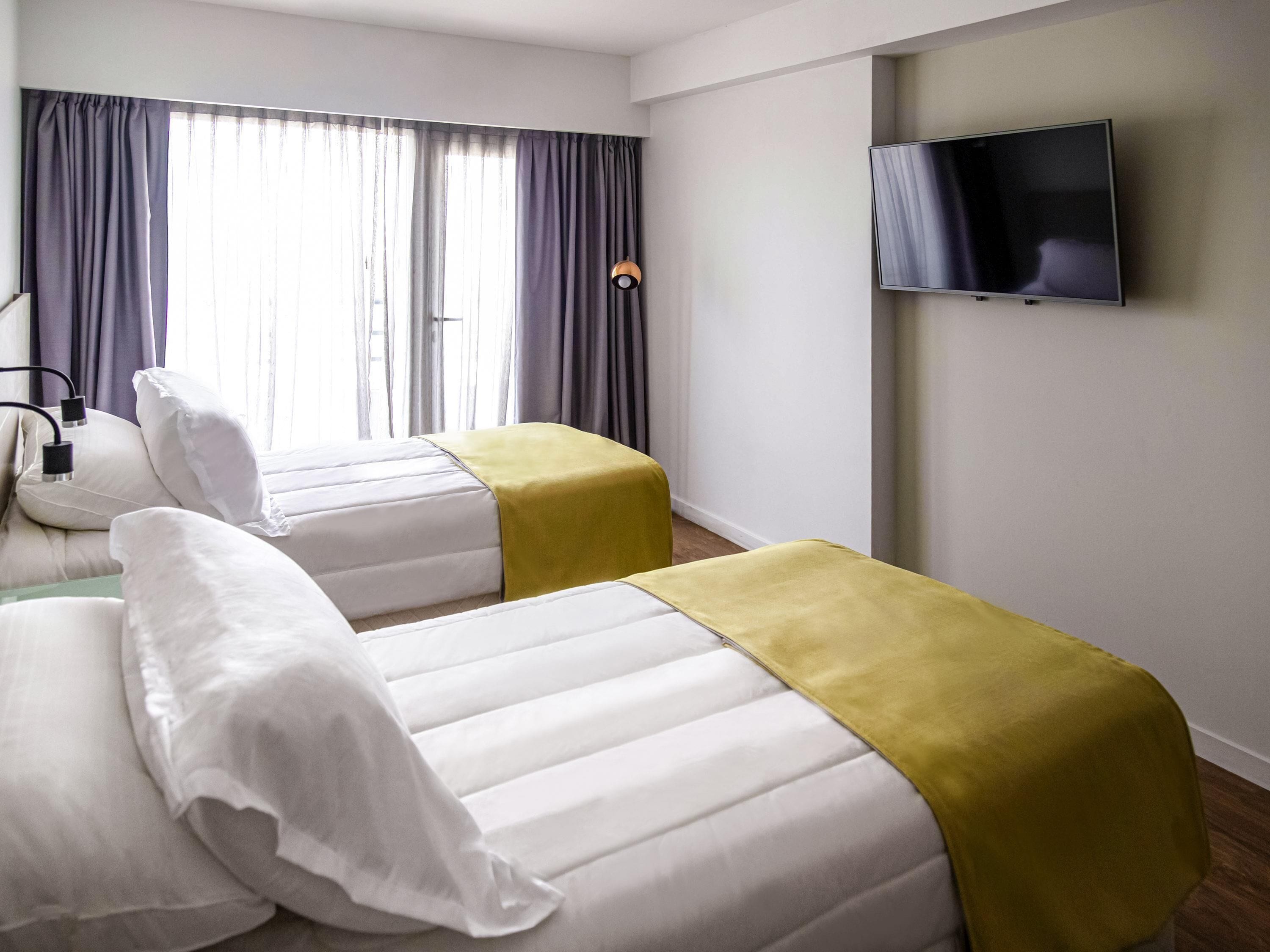 Une chambre d’hôtel avec deux lits, un téléviseur fixé au mur et une fenêtre avec des rideaux.