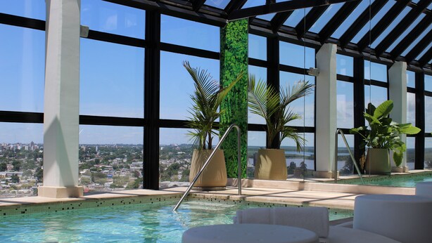 An indoor swimming pool with clear blue water, surrounded by white lounge chairs and large potted plants.
