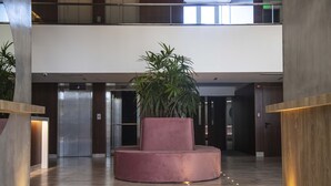A modern lobby with a central circular seating area, a large potted plant, and a pair of elevators.