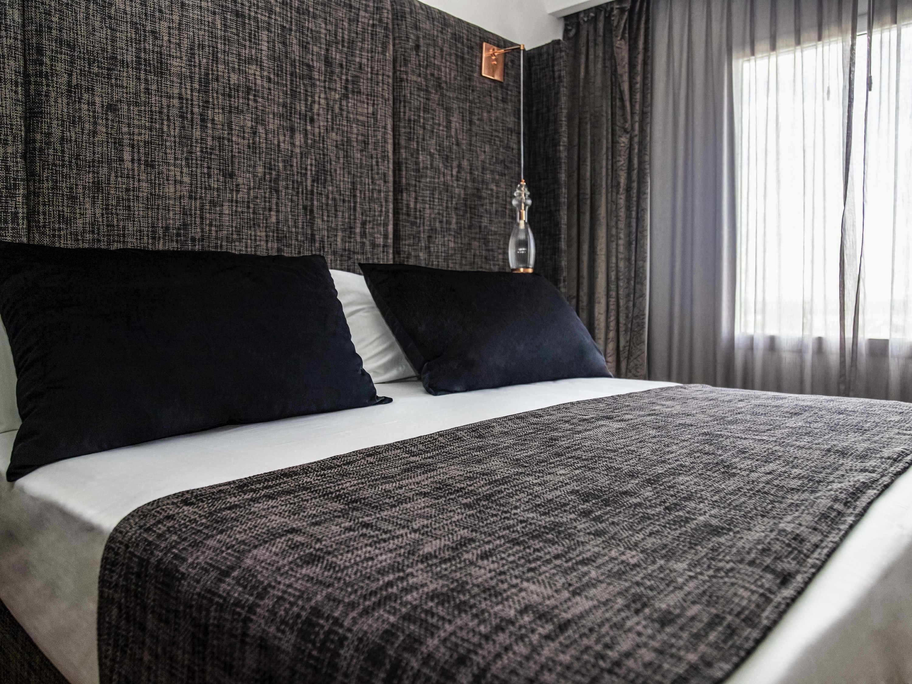 Una cama bien hecha con un edredón oscuro y almohadas, una pared texturizada al fondo y una lámpara colgante.