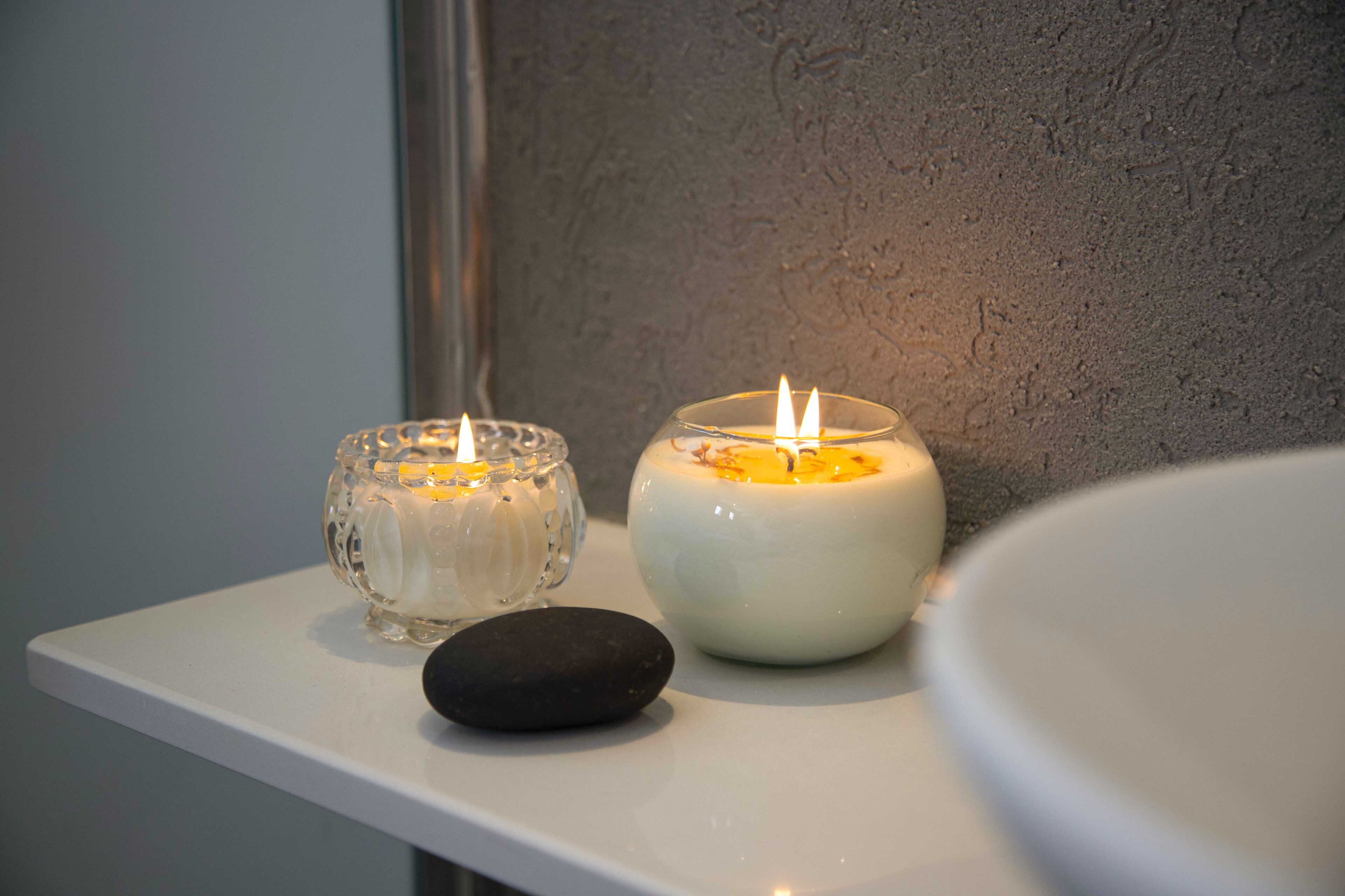 Two lit candles on a white shelf with a stone and a mirror in a bathroom.