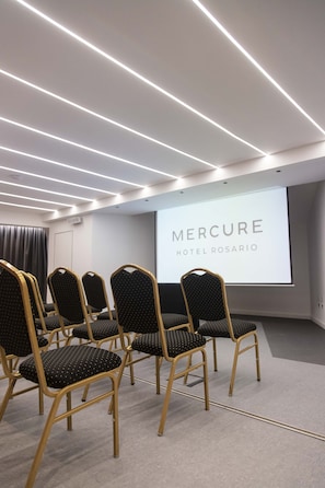 Een vergaderzaal met rijen stoelen, een projectiescherm waarop 'Mercure Hotel Rosario' staat en moderne verlichtingsarmaturen.