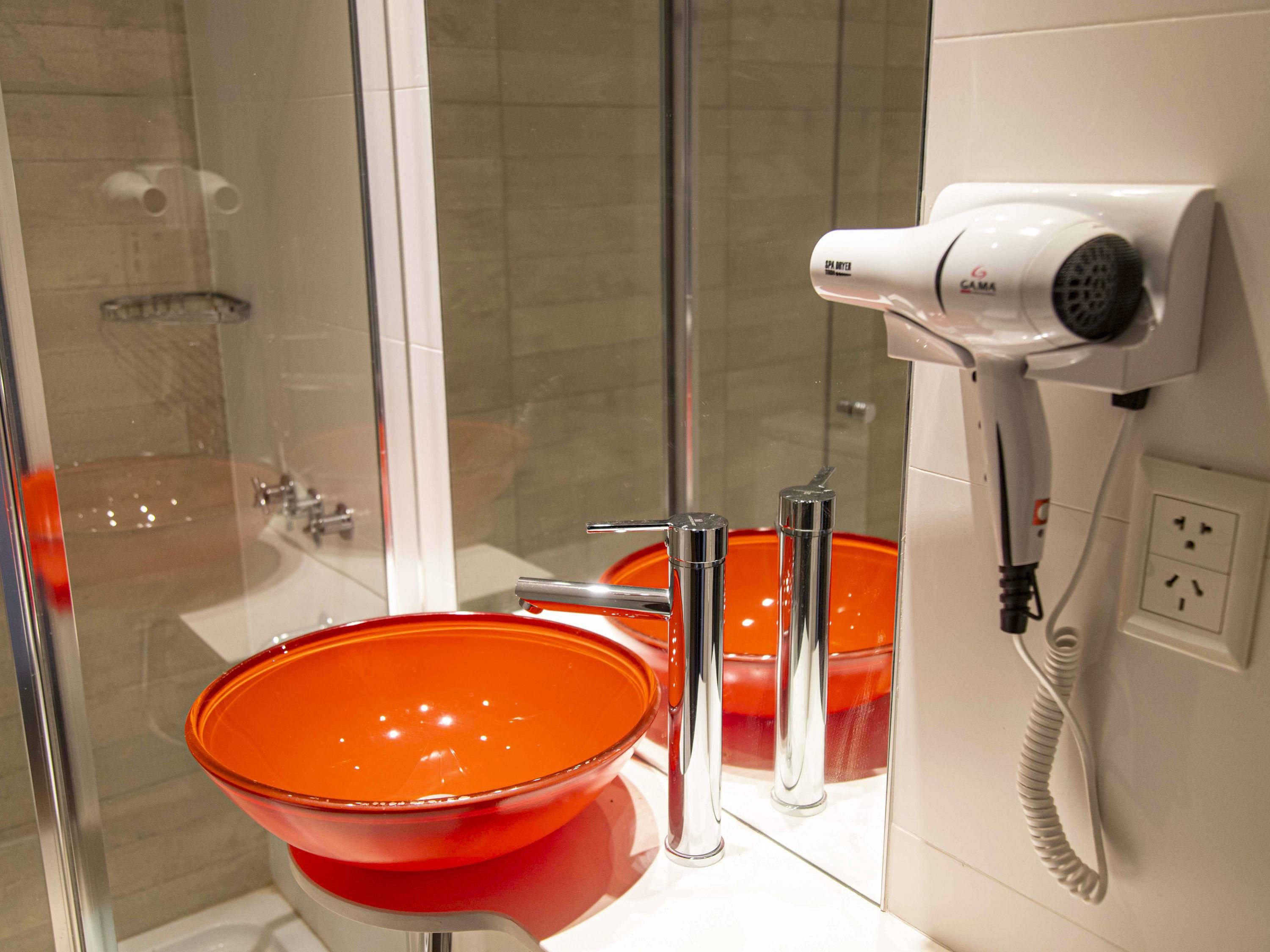 Een badkamer met een douchecabine van glas, een witte haardroger en een oranje wastafel.