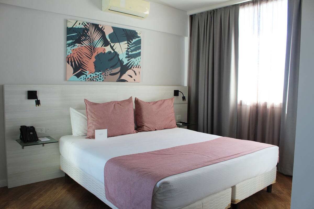Een netjes opgemaakt bed met een witroze sprei, een nachtkastje met een telefoon en een lamp, en een kunstwerk aan de muur boven het bed.