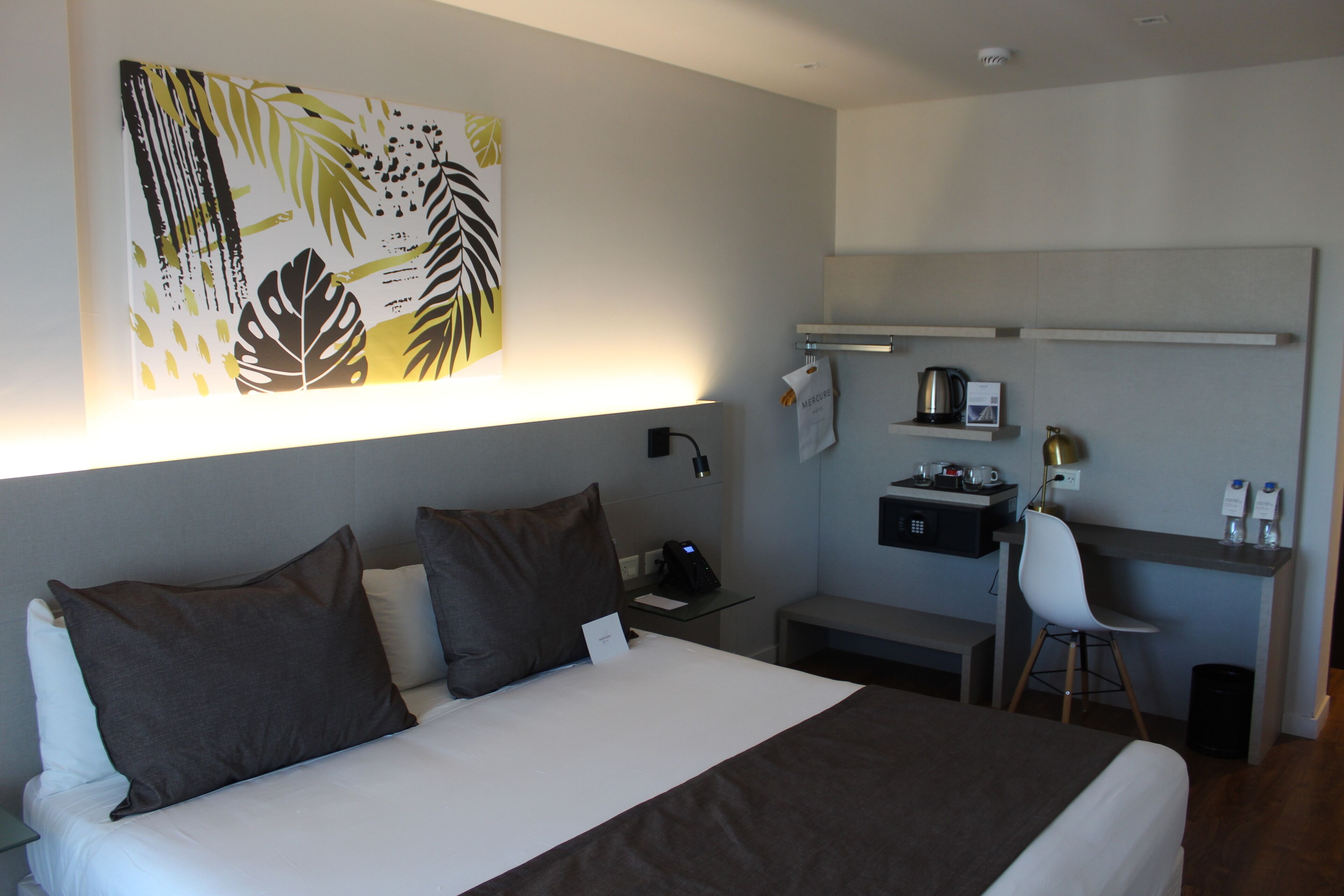 Habitación de hotel con cama, escritorio, silla y estante de pared con una tetera y otros objetos.