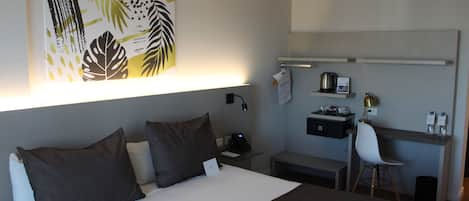 Habitación de hotel con cama, escritorio, silla y estante de pared con una tetera y otros objetos.
