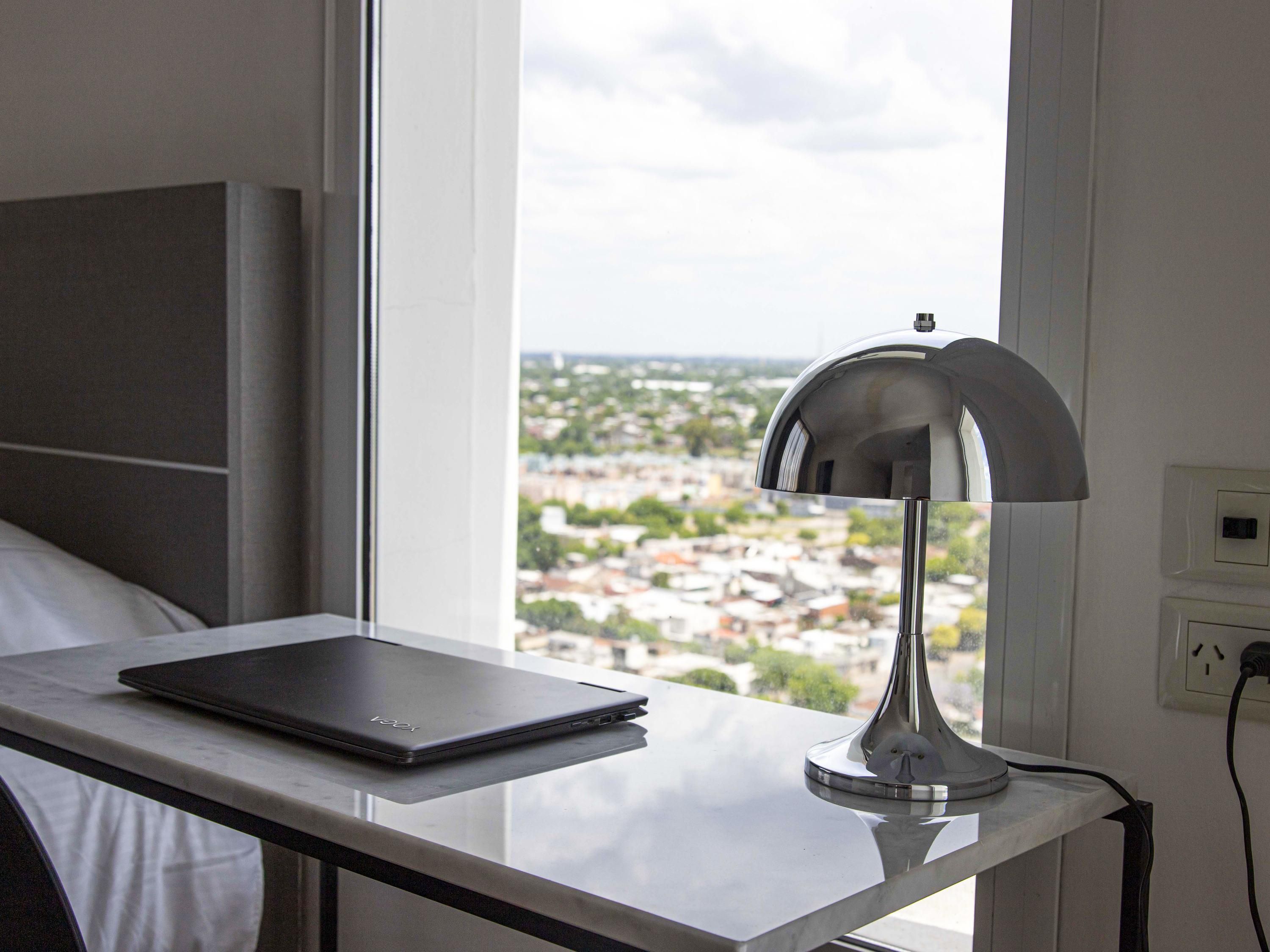 A laptop on a table near a window with a view of a cityscape.
