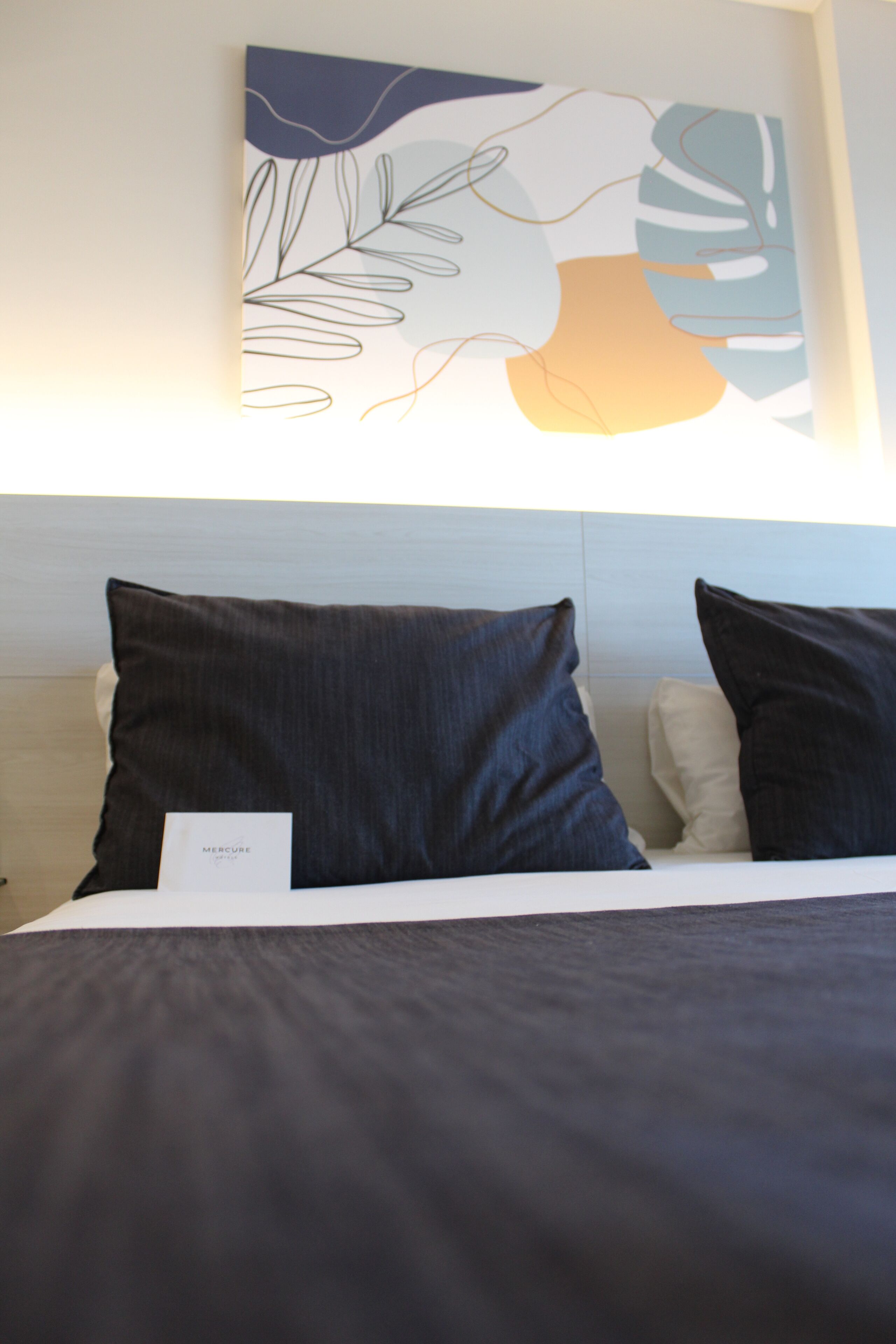 A neatly made bed with dark bedding and two pillows, a wall art piece above, and a white headboard.