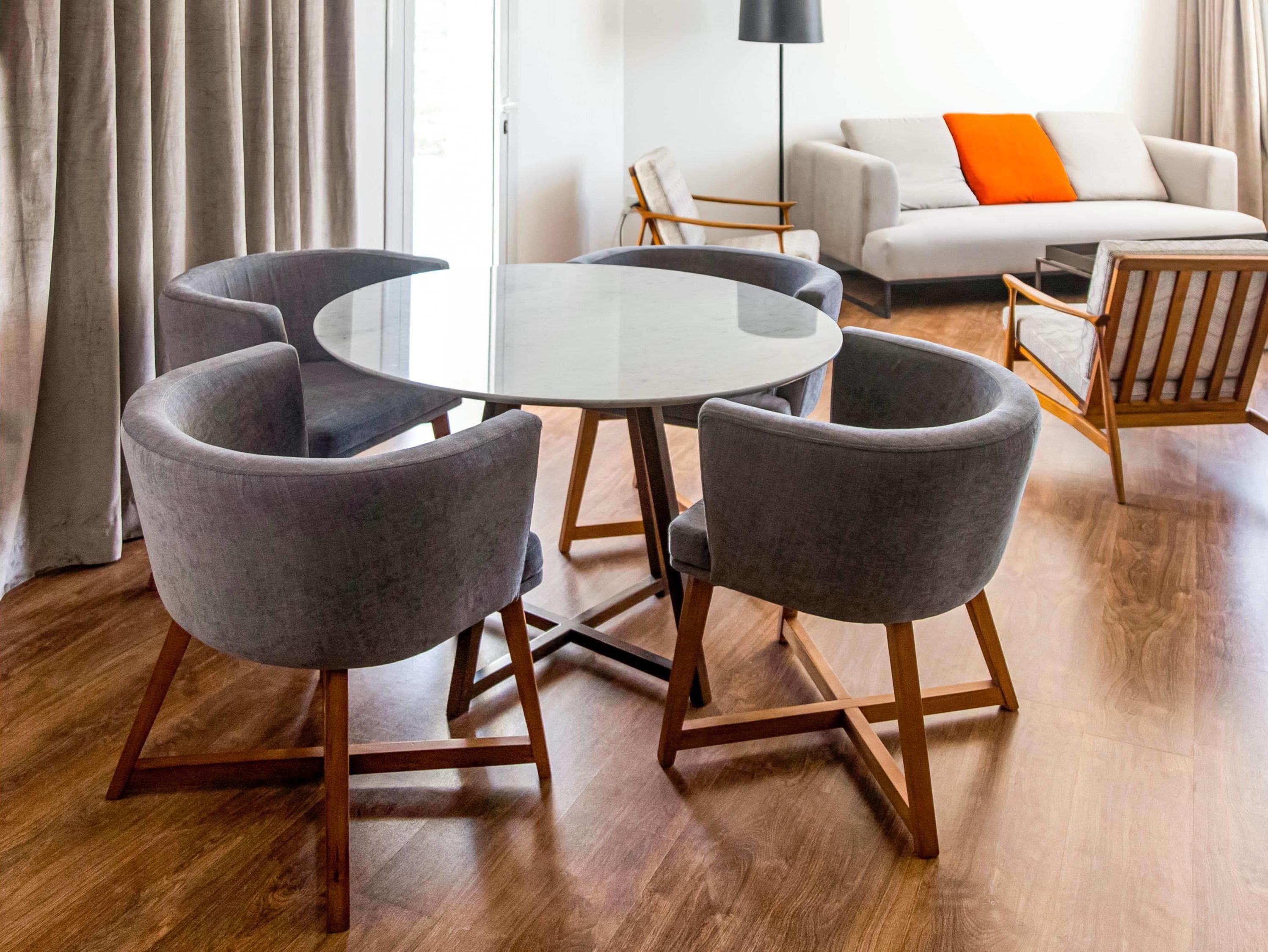 A modern living room with a round glass table surrounded by four grey armchairs and a wooden floor.