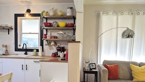 Cocina moderna con armarios blancos, un lavamanos y una ventana sobre la encimera. Hay un comedor con una mesa y sillas, y una sala de estar con un sofá y una lámpara.