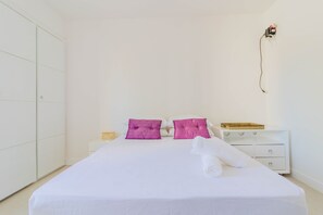 Cama bem arrumada com roupa de cama branca e duas almofadas roxas, mesa de cabeceira branca com bandeja e guarda-roupa branco.