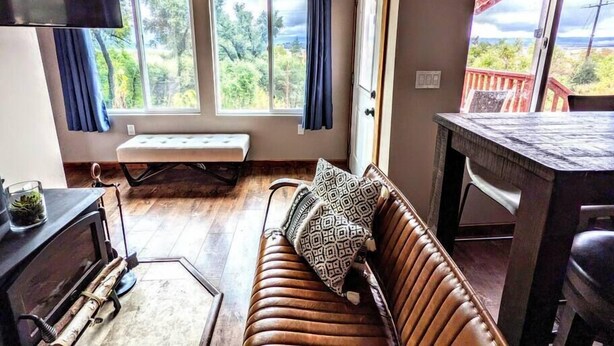 A cozy room with a brown leather sofa, a bench, and a dining table. Large windows offer a view of trees and a red deck outside.