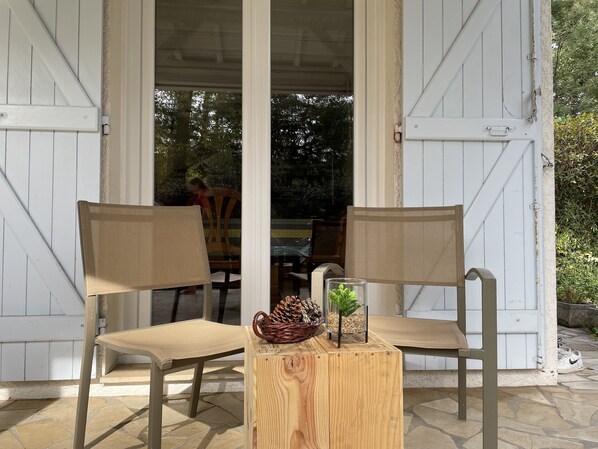 Two chairs with a woven basket and a glass container with a plant on a wooden table outside a building with white doors. - A cozy house in a central location on the Cap Ferret peninsula (Lège-Cap-Ferret)