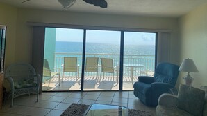 A room with a ceiling fan, a large sliding glass door leading to a balcony with chairs and a view of the ocean.