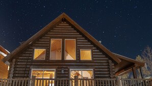 Una cabaña de troncos con balcón y ventanas amplias, con un cielo estrellado de fondo.