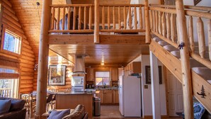 Interior de una cabaña de troncos con comedor, cocina y una escalera de madera.
