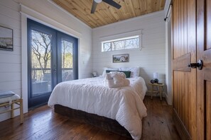 A bedroom with a large bed, wooden flooring, a ceiling fan, a window with a view, and a door. - Secluded Treehouse Cabin Near Hochatown with Hot Tub, Game Loft & Playset (Broken Bow)