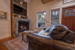 A cozy living room with a stone fireplace, leather sofa, and a large window offering a view of the outdoors. - Secluded Treehouse Cabin Near Hochatown with Hot Tub, Game Loft & Playset (Broken Bow)