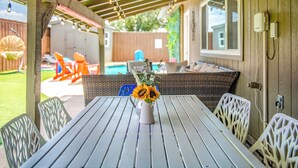 Een overdekt terras met een tafel en stoelen, een vaas met zonnebloemen en uitzicht op een achtertuin met een zwembad en tuinmeubilair.