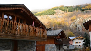 A wooden chalet with a stone facade, a balcony with intricate railings, and a view of a forested hillside. - Le Refuge du Cabri -15 people- sauna - jacuzzi Les Ménuires -Skis aux Pieds (LES BELLEVILLE)
