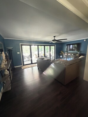 A living room with a dark wood floor, a ceiling fan, a large TV, and a sliding glass door leading to an outdoor patio.