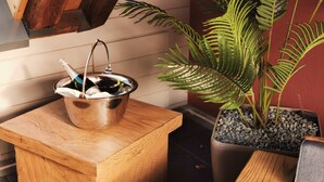 Un seau en métal avec une bouteille posée sur une table en bois, une plante en pot et un mur rouge.