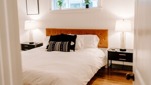 Une chambre à coucher avec un grand lit, deux tables de chevet, une fenêtre ornée de plantes et un tableau encadré accroché au mur.