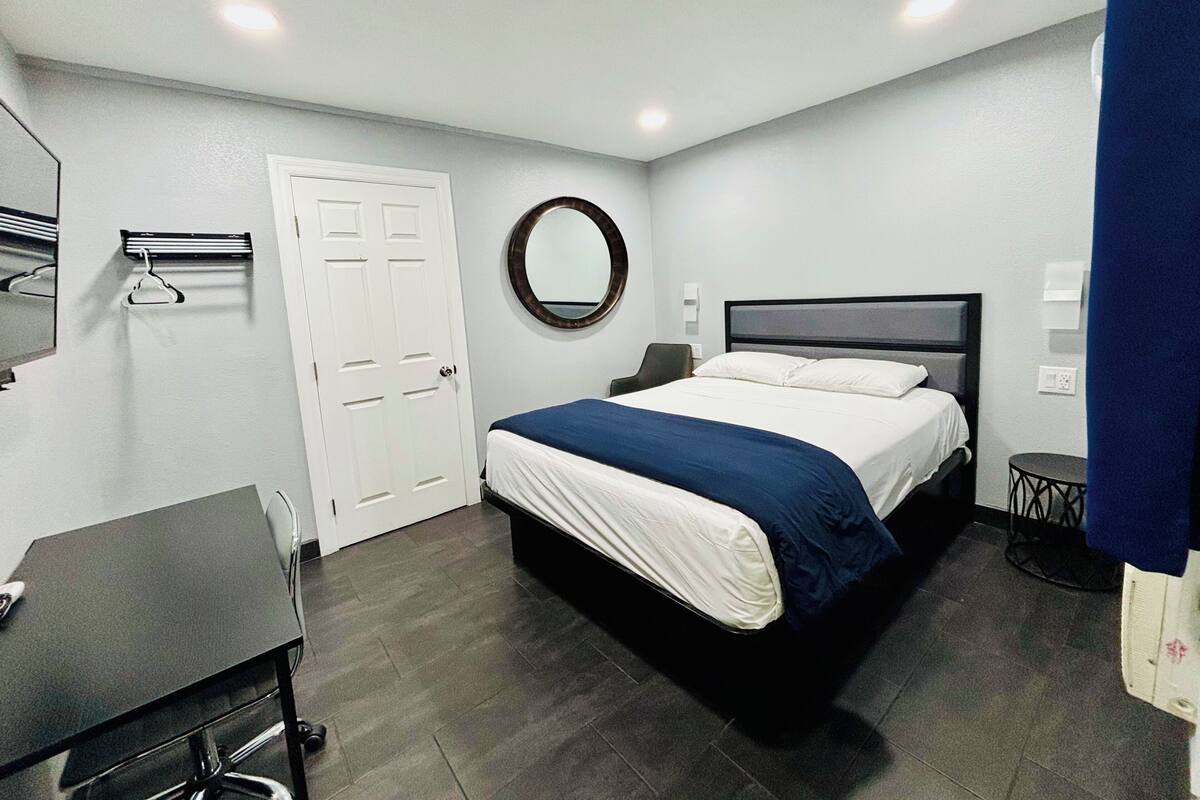 A hotel room with a bed, a desk, a chair, a round mirror, and a door.