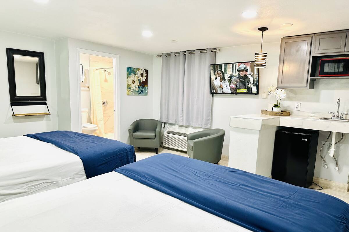 A hotel room with two beds, a desk, a chair, a TV, and a kitchenette.