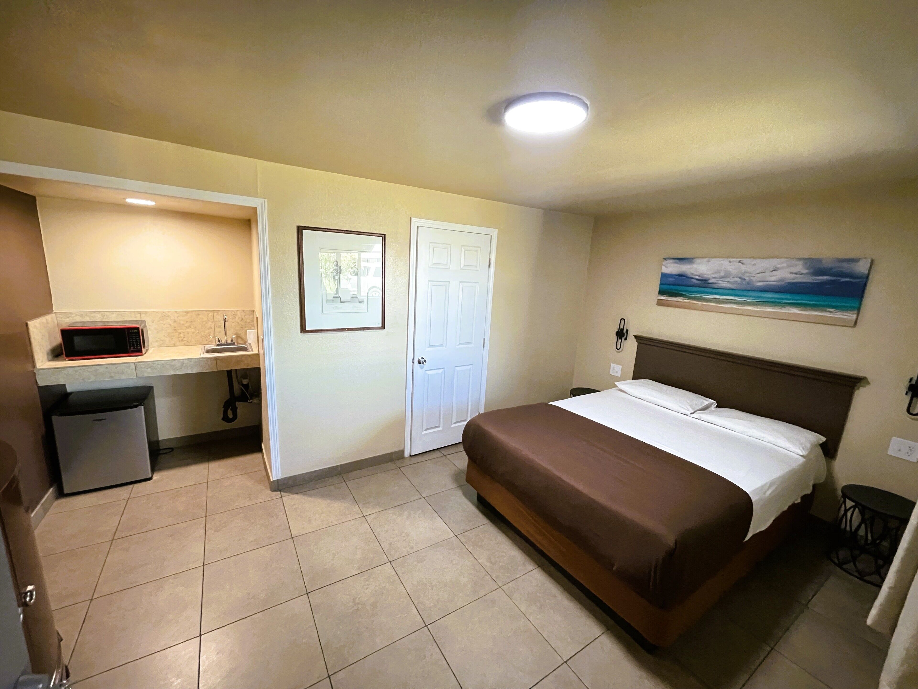 A hotel room with a bed, a small kitchenette, a bathroom, and a painting on the wall.