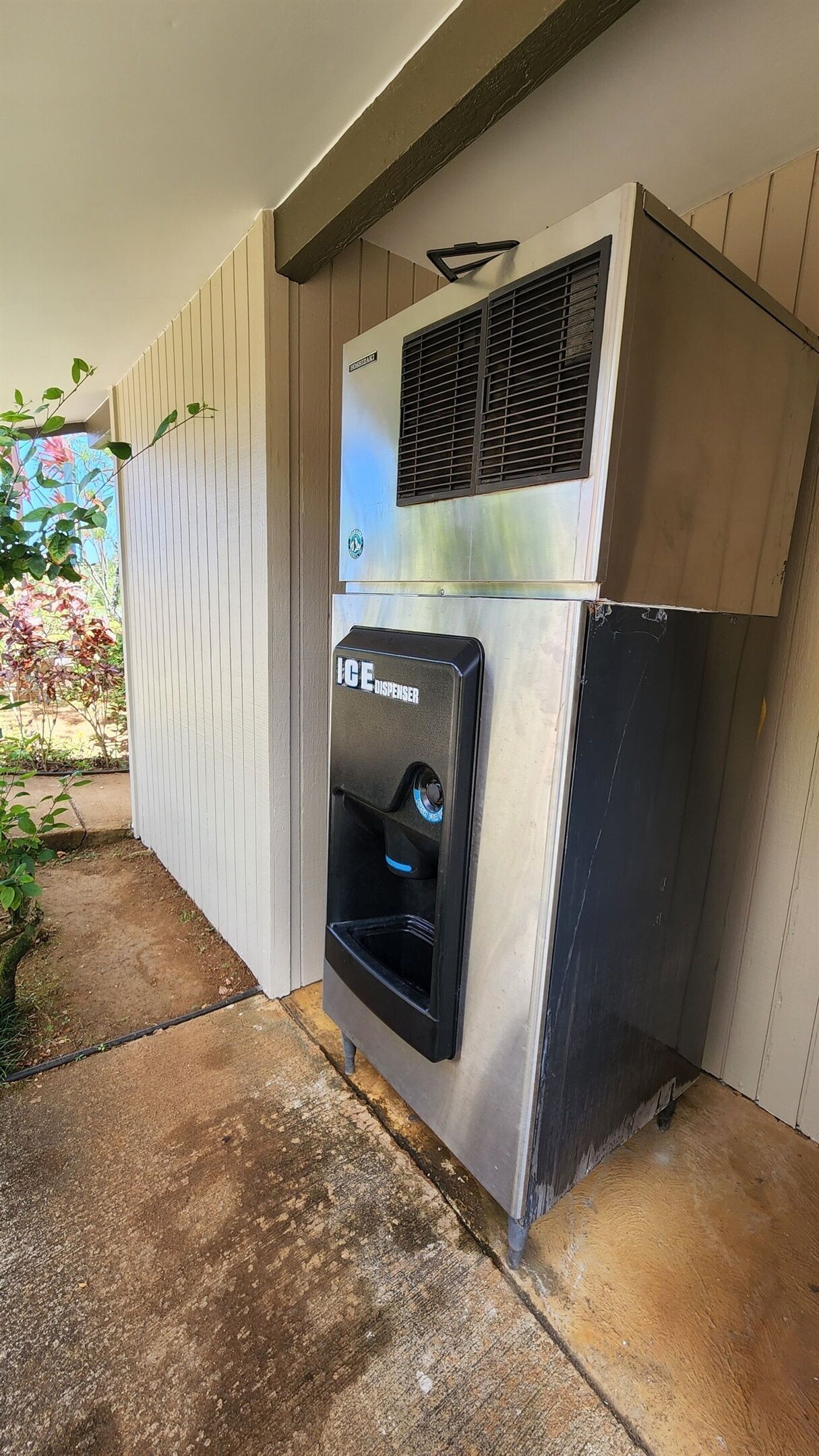 A portable ice dispenser attached to a metal cabinet, located outdoors against a wooden wall.