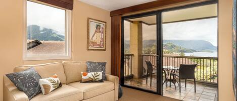 A room with a beige sofa, a painting, and a view of a balcony with chairs and a table.