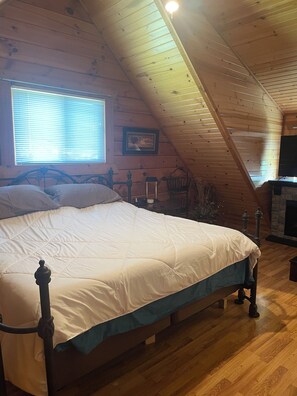 A bedroom with a bed, wooden ceiling, a window with blinds, a framed picture on the wall, and a fireplace.