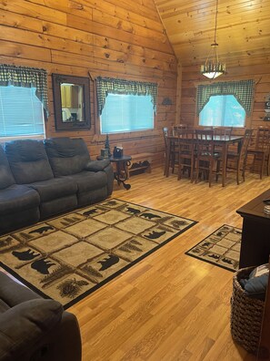 A cozy cabin interior with a wooden ceiling, a dining table with chairs, a sofa, and a small Christmas tree.