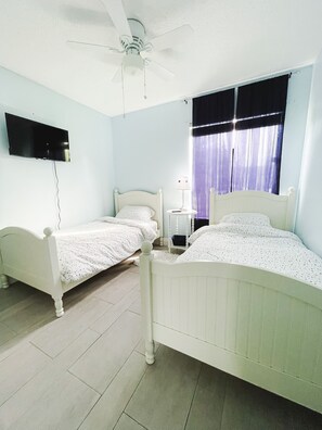 A bedroom with two single beds, a ceiling fan, a TV mounted on the wall, and a window with curtains.