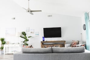 A modern living room with a grey sofa, a wooden coffee table, and a ceiling fan. There are wall-mounted artworks and a flat-screen TV. - Waterfront Boca Raton Home - Pets OK - Heated Pool (Boca Raton)