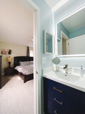 A bedroom with a bed, bedside table, lamp, and a large mirror above a white sink with a blue cabinet below.