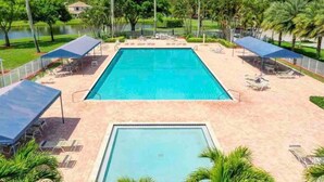 Una piscina rectangular con una terraza alrededor, protegida por sombrillas, rodeada de palmeras y una extensión de agua.