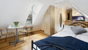 Une chambre avec un plafond en pente, un lit recouvert d’une couverture bleue, une armoire en bois, une petite table ronde avec des chaises et une fenêtre donnant sur un immeuble.